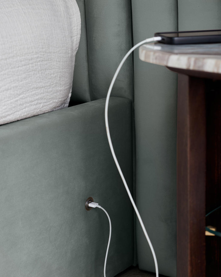 Bedside table with a phone plugged into the bed. Plush headboard and a modern style.