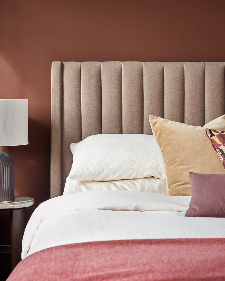 A stylish bedroom setup featuring a headboard, bedding, pillows, a side table and a lamp.