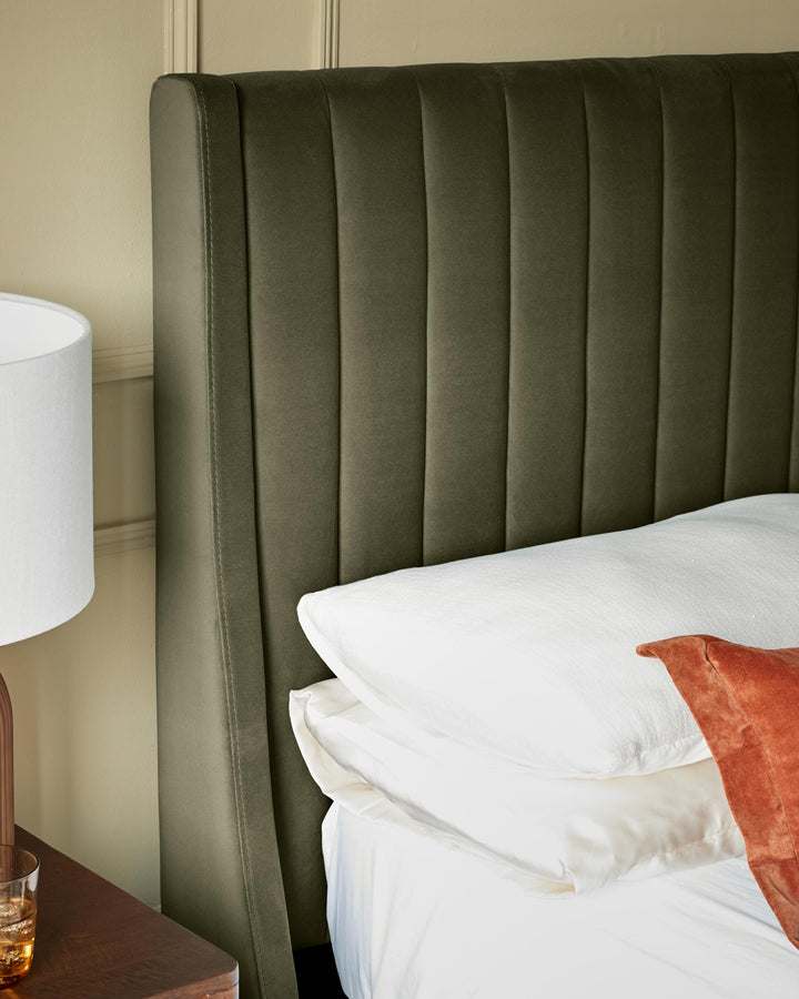 An olive green, velvet, fluted headboard sits behind a stack of white pillows on a bed, next to a lamp and wooden table.
