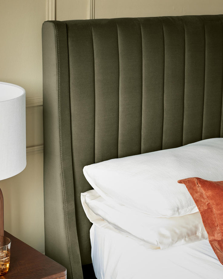 An olive green, velvet, fluted headboard sits behind a stack of white pillows on a bed, next to a lamp and wooden table.