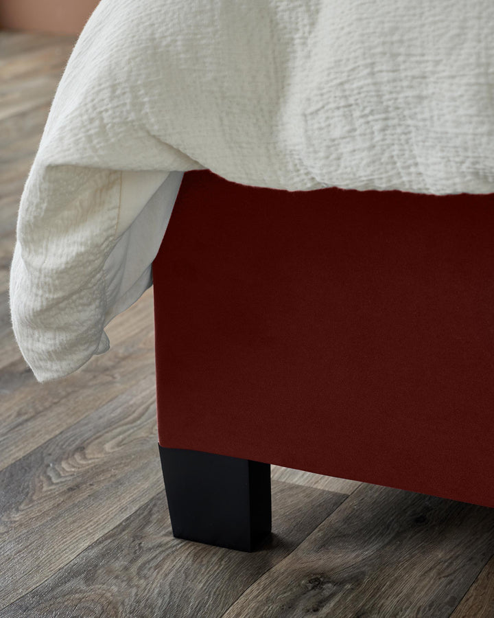 A bed frame with a red fabric finish and black legs, partially covered with a white duvet cover.