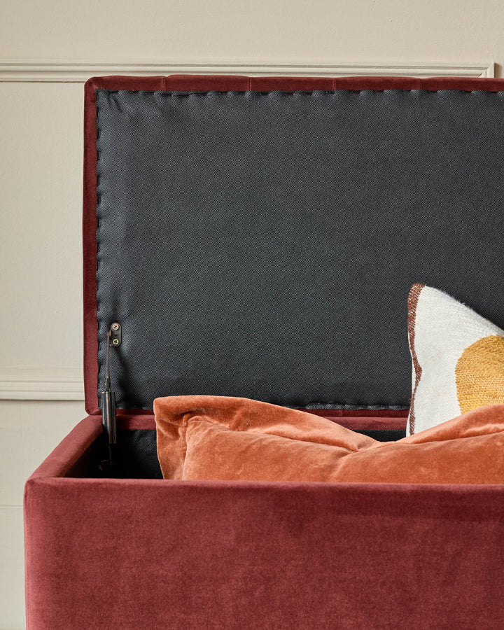 Toffee-red velvet ottoman bench with open storage lid, revealing dark interior, styled with decorative pillows.