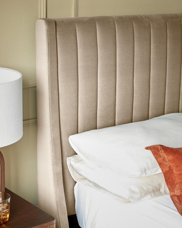 Close-up of a stylish bed with a tall, ribbed headboard, pillows, and a wooden bedside table with a lamp.
