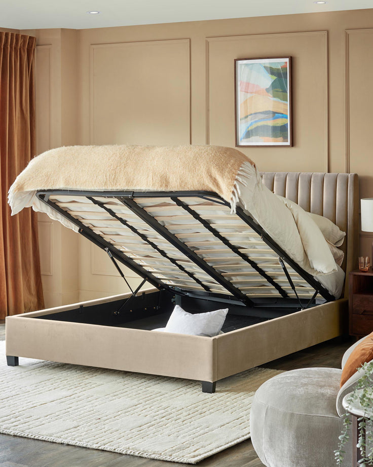 A beige ottoman bed with lift-up frame and storage, set in a neutral room with a velvet chair and abstract art.