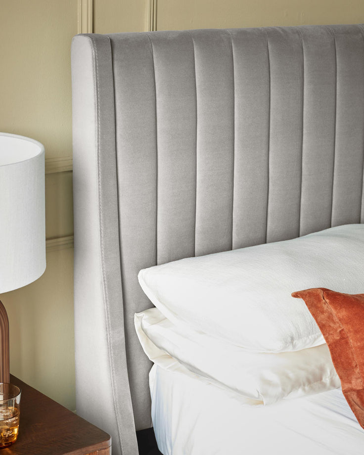 A luxurious bed with a vertically channelled grey headboard, crisp white linens and a dark wood bedside table.
