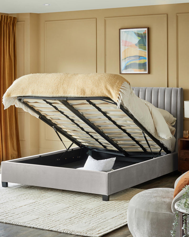 A luxurious grey ottoman bed with headboard lifted to reveal storage space, on a textured rug in a neutral toned room.