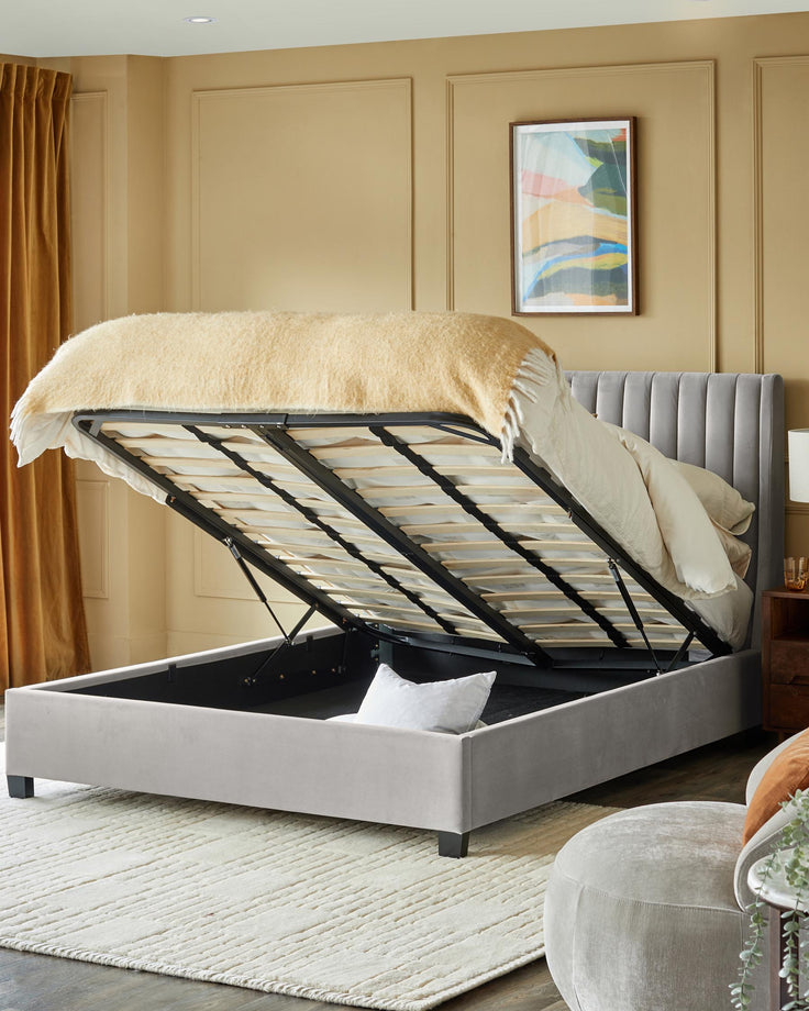 A luxurious grey ottoman bed with headboard lifted to reveal storage space, on a textured rug in a neutral toned room.