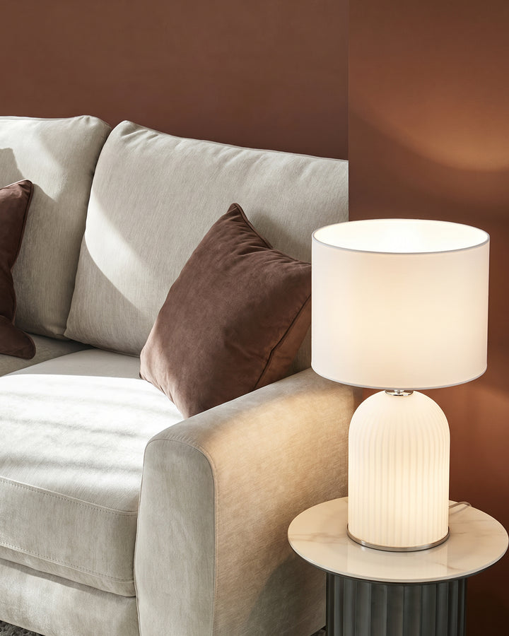 Amalfi white table lamp with ribbed ceramic base and cylindrical fabric shade, displayed on marble side table next to cream sofa.