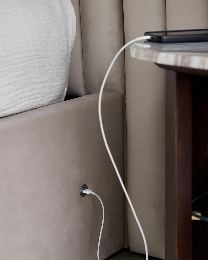 A tan upholstered headboard with a dark wood nightstand. A phone charges on the table.