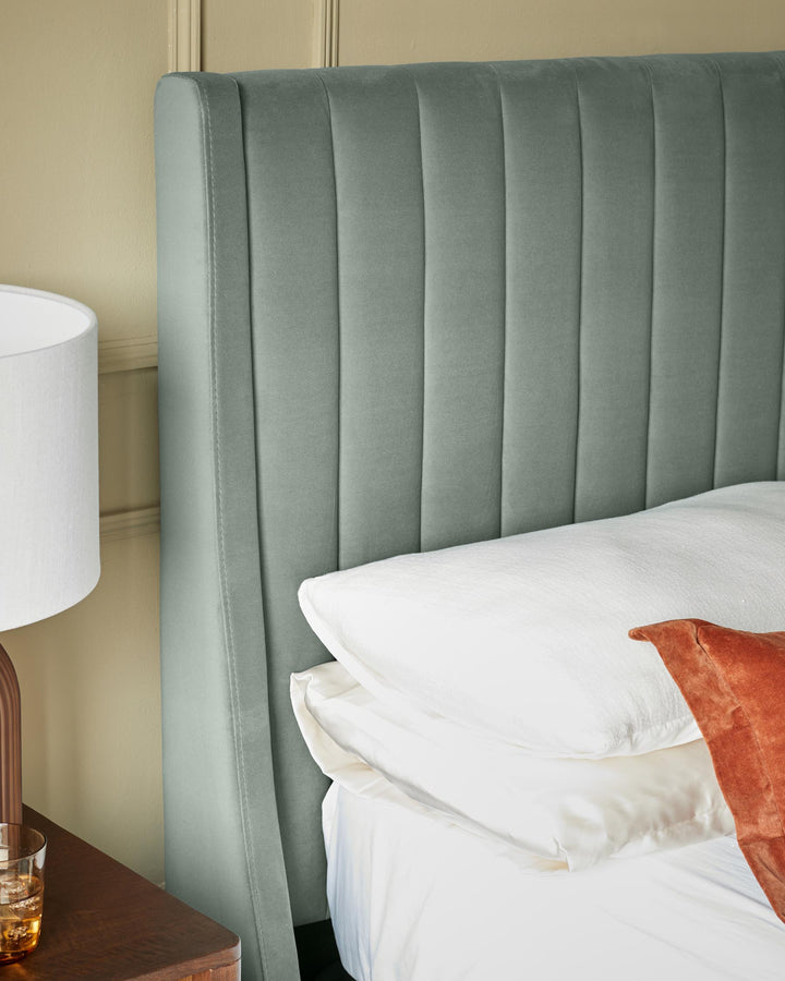 Close up of a plush upholstered bedhead, side table with lamp and drink, and bedding with pillows
