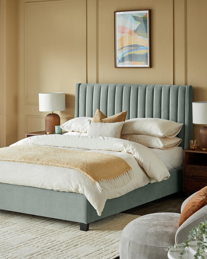 Bedroom with a blue fabric bed, bedside tables, lamps, and a rug. Artwork hangs on the wall above the bed.