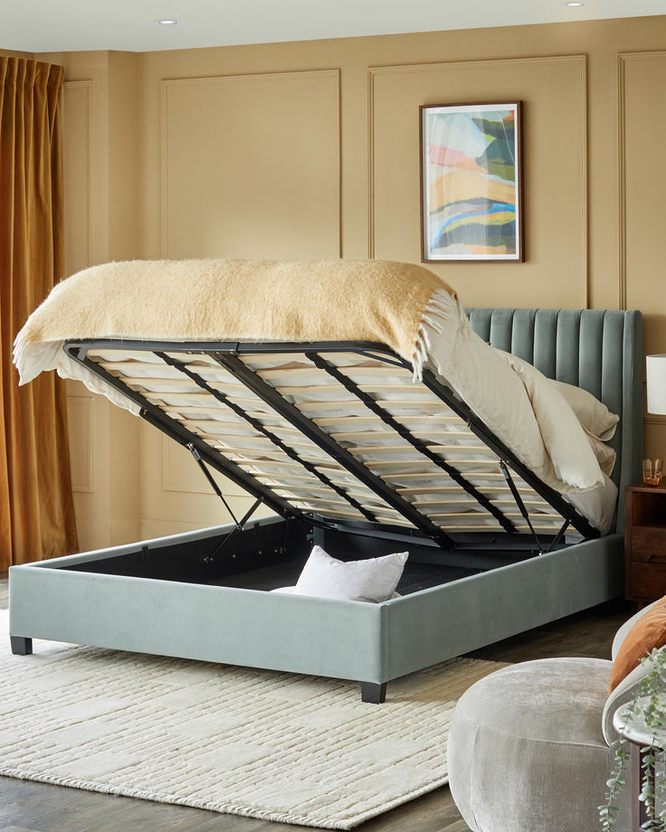 Upholstered ottoman storage bed in light green fabric, partially open to reveal hidden space. Styled with rug and side table.