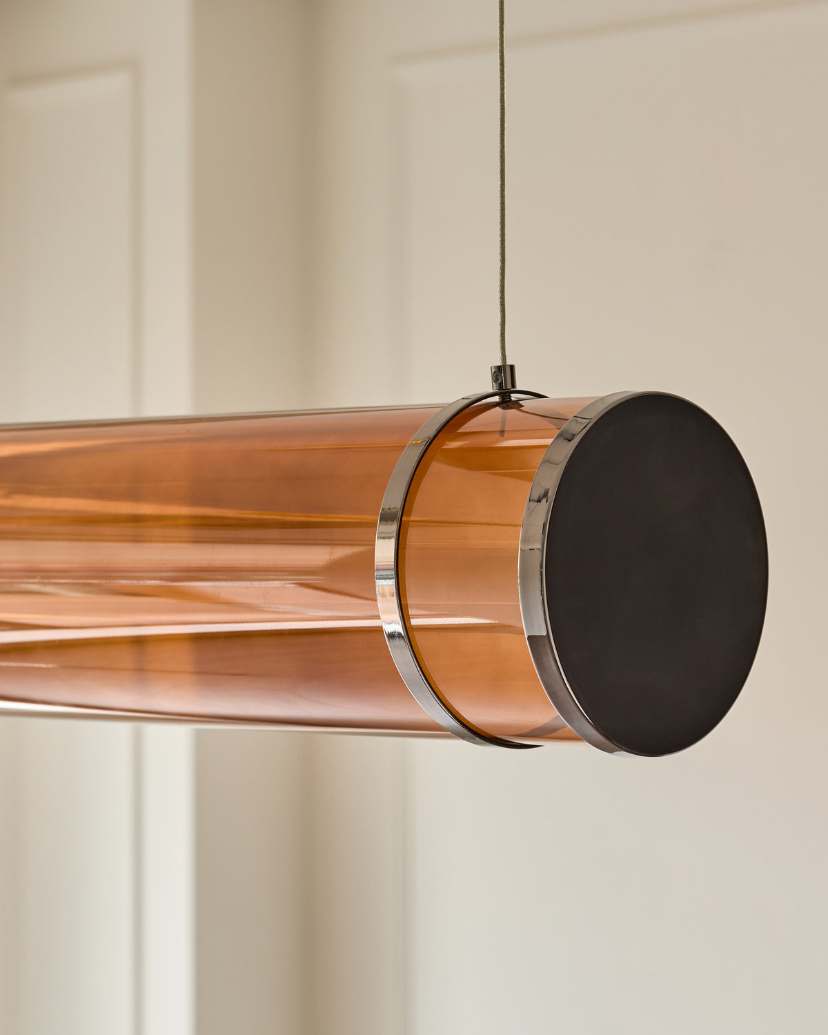 Amber glass pendant light with caramel finish, cylindrical shape, chrome bands, and black circular end cap, hanging from cord.