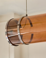 Amber single pendant light with caramel glass cylinder, chrome metal bands, suspended by cord against neutral background.
