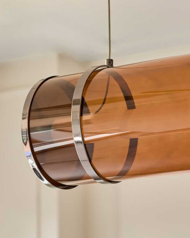 Amber single pendant light with caramel glass cylinder, chrome metal bands, suspended by cord against neutral background.