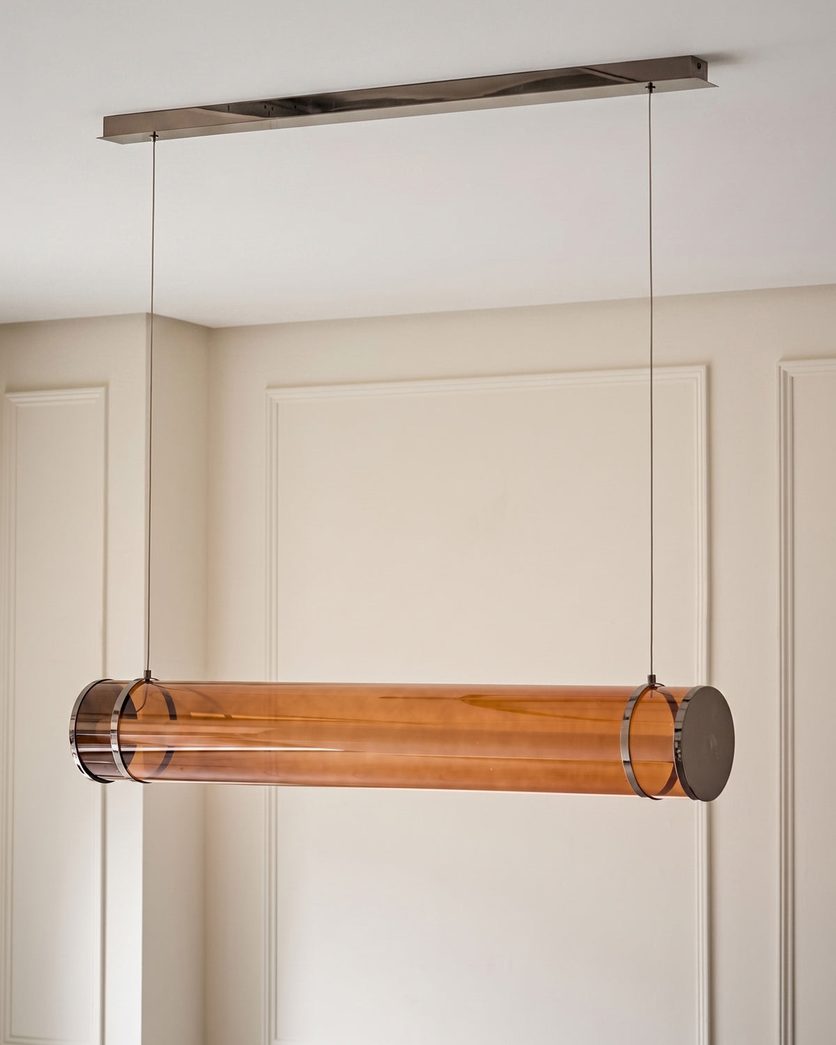 Amber glass pendant light with caramel-tinted cylindrical shade, suspended from brushed metal ceiling canopy by thin cables.