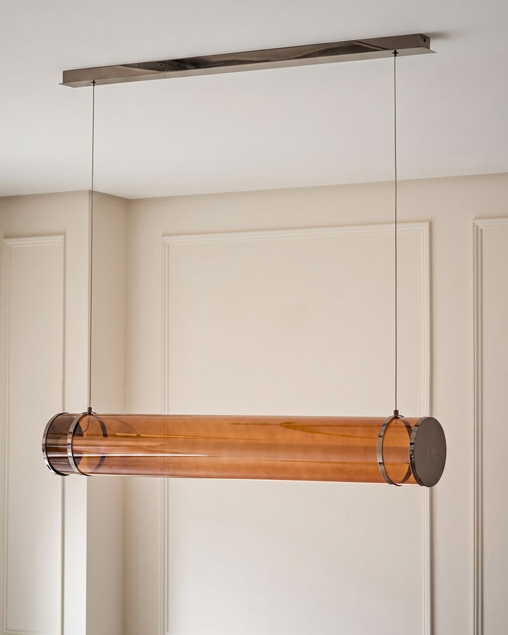 Amber glass pendant light with caramel-tinted cylindrical shade, suspended from brushed metal ceiling canopy by thin cables.