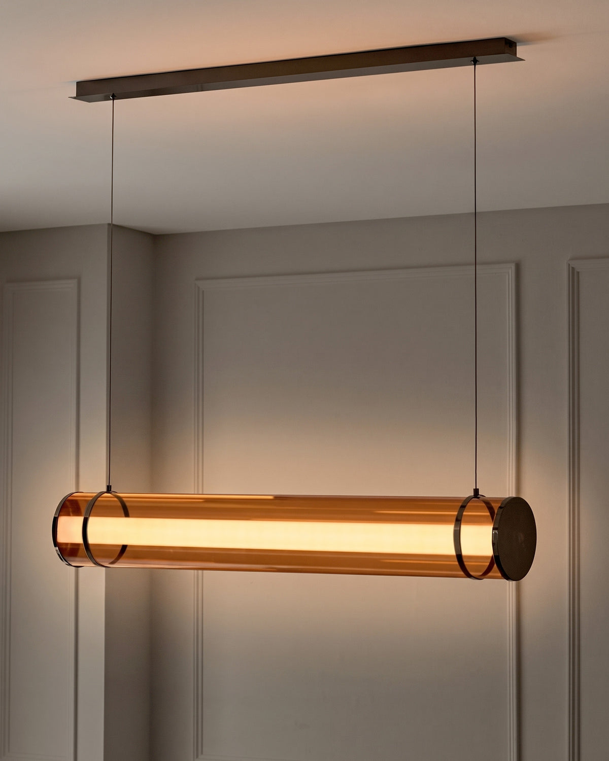 Amber glass pendant light with caramel-toned cylindrical shade, suspended from dark metal ceiling fixture with chain hardware.