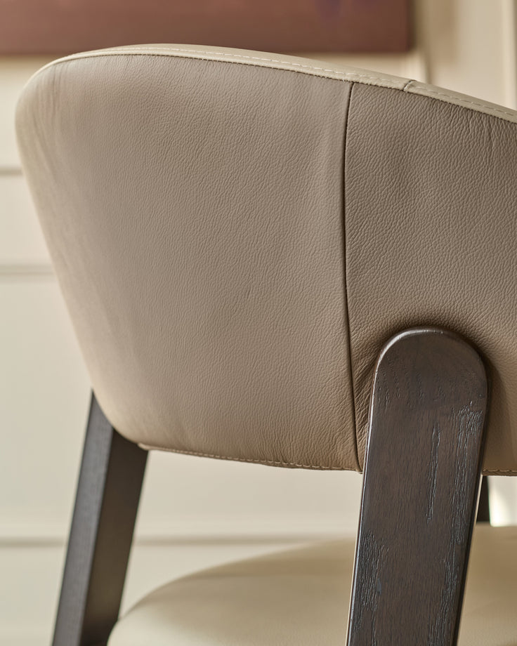 Amelia dining chair in natural leather with mocha wood legs, detail view of curved backrest and wooden frame.