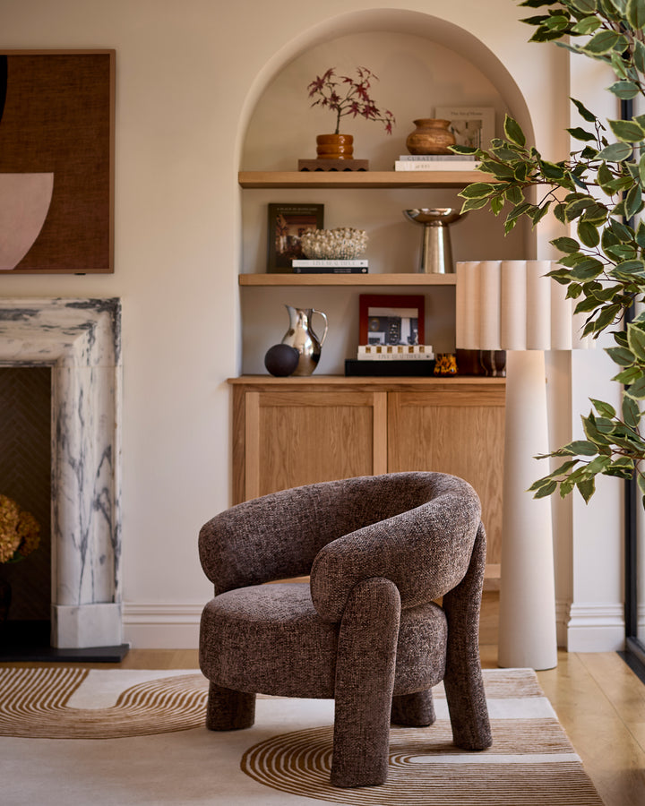 Mocha fabric accent chair with curved backrest and sturdy wooden legs, displayed in a bright living room setting.
