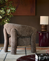 Anya accent chair in mocha fabric with cylindrical legs, displayed in a styled living room setting with a burgundy side table and lamp.