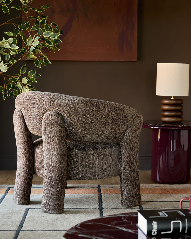 Anya accent chair in mocha fabric with cylindrical legs, displayed in a styled living room setting with a burgundy side table and lamp.
