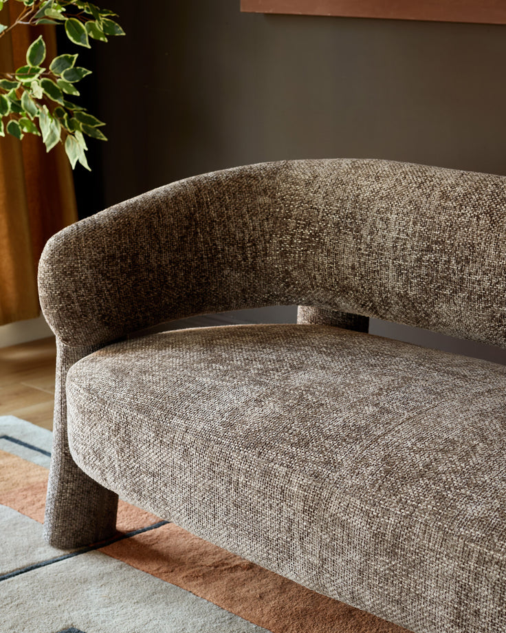 Mocha fabric accent sofa with curved back and cylindrical legs, angled side view on geometric area rug.