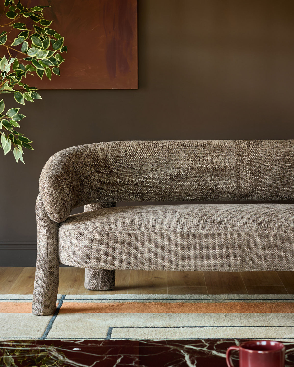 Anya accent sofa in mocha fabric, shown from side angle with curved backrest and cylindrical legs on geometric patterned rug.