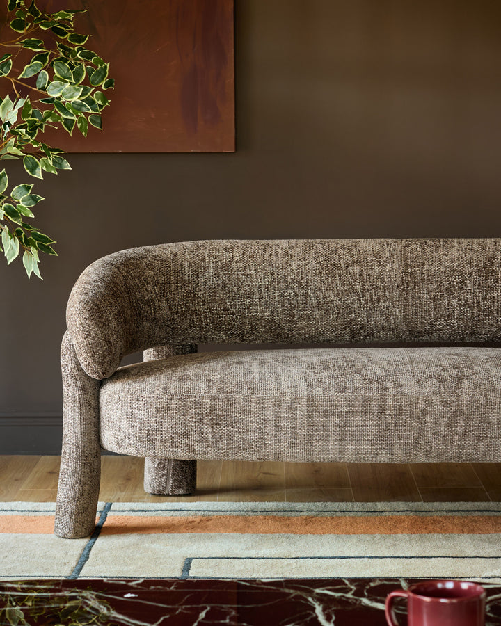 Anya accent sofa in mocha fabric, shown from side angle with curved backrest and cylindrical legs on geometric patterned rug.