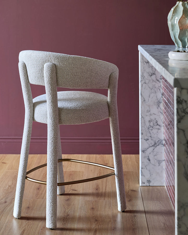 Anya bar stool in natural fabric with curved backrest, cylindrical legs, and dark metal footrest against burgundy wall.