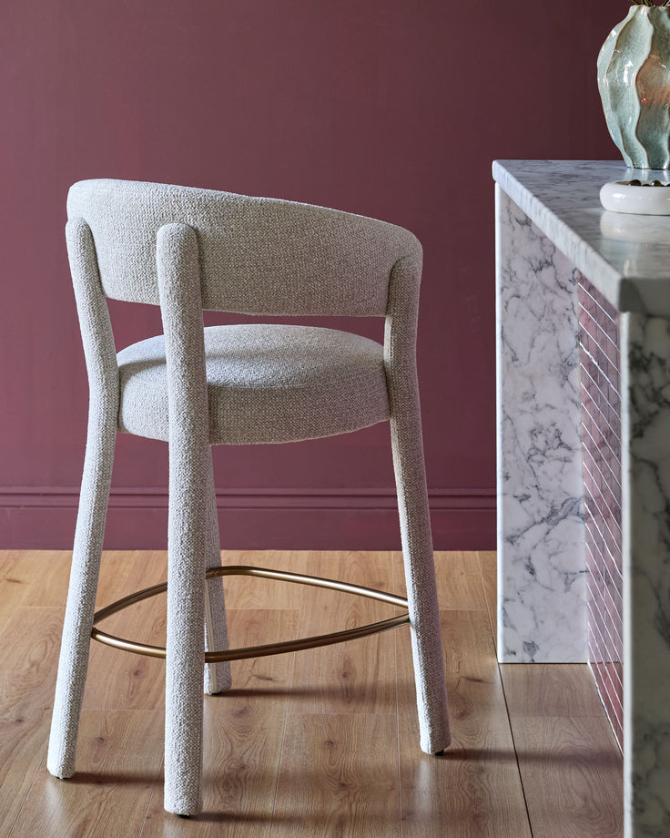 Anya bar stool in natural fabric with curved backrest, cylindrical legs, and dark metal footrest against burgundy wall.