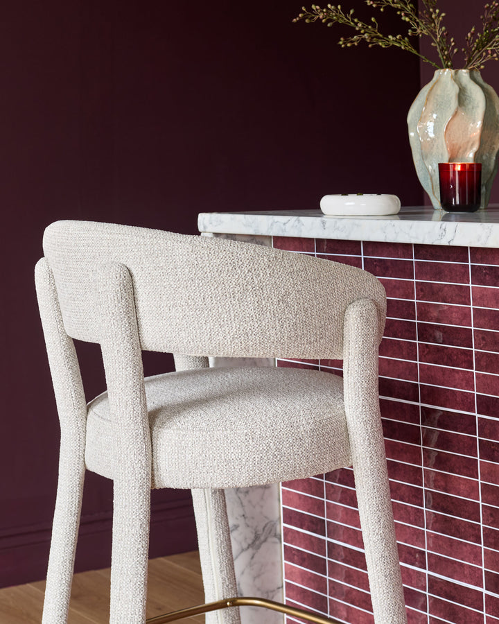 Anya bar stool in natural fabric, shown from side angle at marble counter with burgundy tile backsplash and dark wall backdrop.