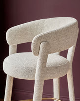 Anya bar stool in natural fabric with curved backrest and armrests, shown at an angle against a burgundy background.