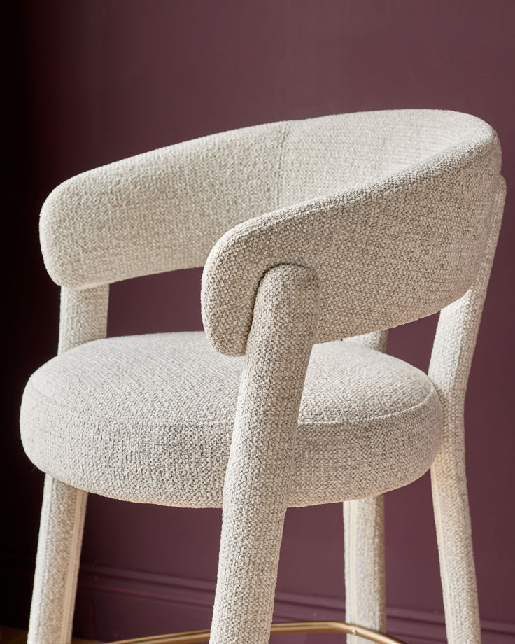 Anya bar stool in natural fabric with curved backrest and armrests, shown at an angle against a burgundy background.