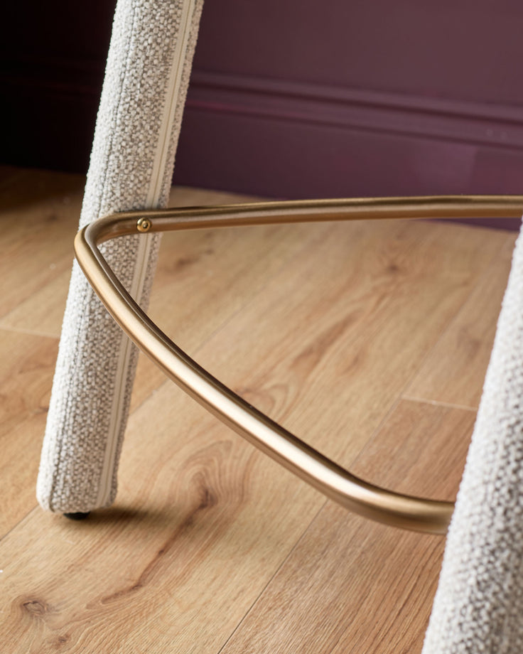 Anya bar stool in natural fabric with brass curved footrest, shown at an angle on wooden flooring.