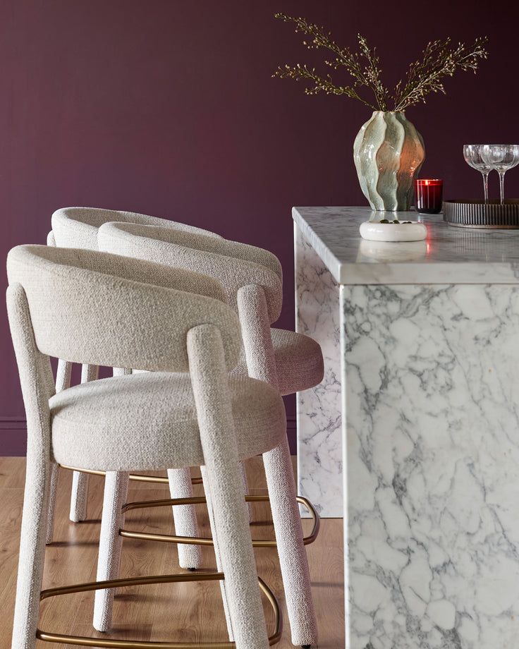 Anya bar stool in natural fabric, shown from back angle with brass footrest, paired with marble counter.