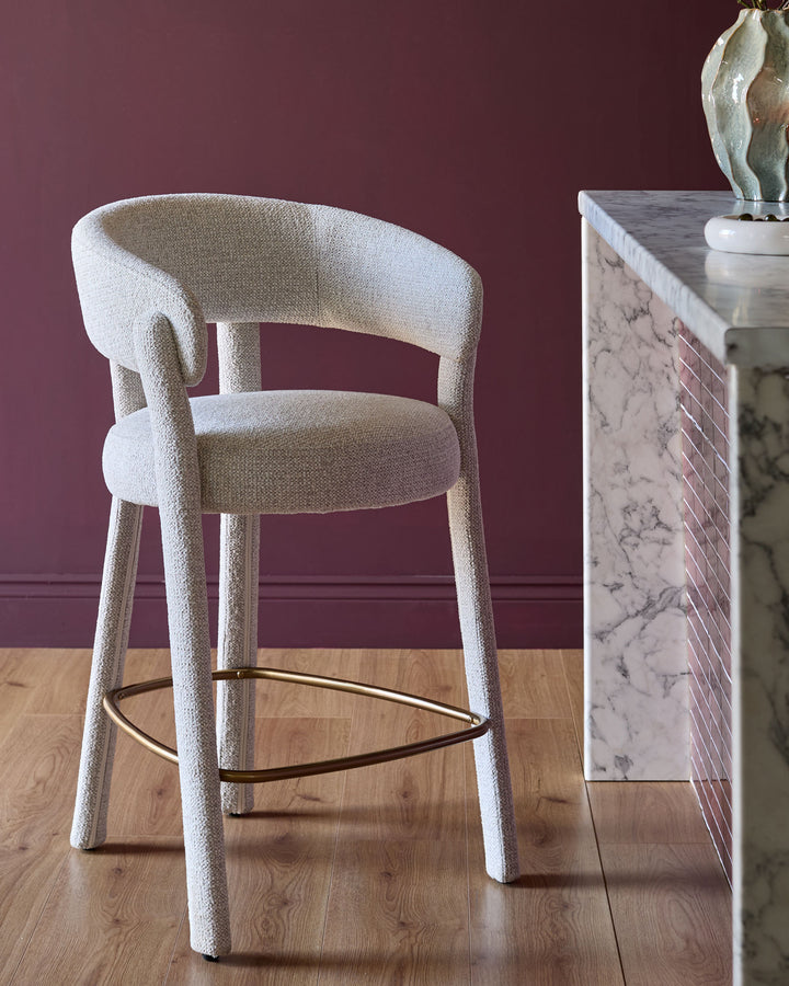Anya bar stool in natural fabric with curved back, brass footrest, and tapered legs against burgundy wall.