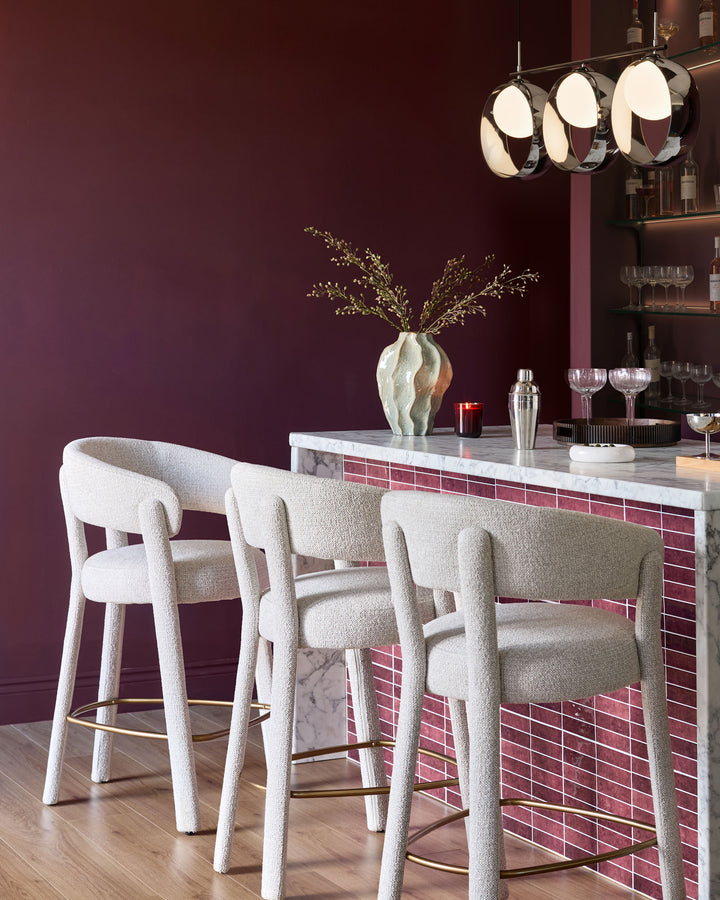 Anya bar stool in natural fabric with curved backrest and brass footrest, shown in burgundy bar setting.