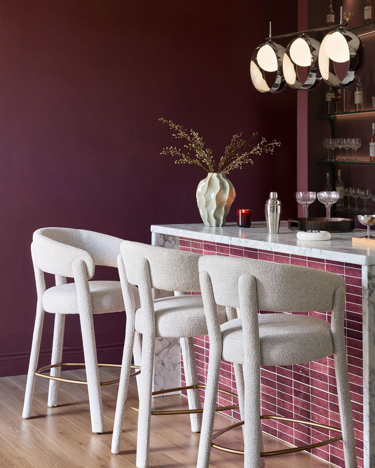 Anya bar stool in natural fabric with curved backrest and brass footrest, shown in burgundy bar setting.