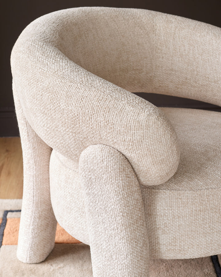 Anya accent chair in natural fabric, curved silhouette with rounded armrests and cylindrical legs, side angle view.