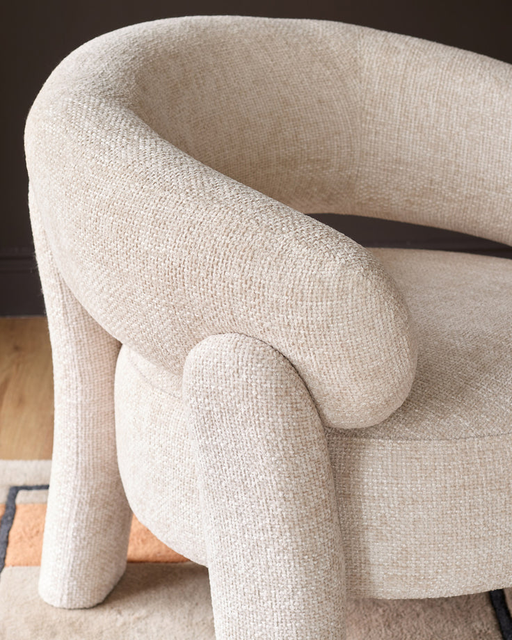 Anya accent chair in natural fabric, curved silhouette with rounded armrests and cylindrical legs, side angle view.