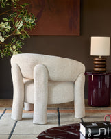 Anya accent chair in natural fabric with curved arms and cylindrical legs, displayed in a styled living room setting.