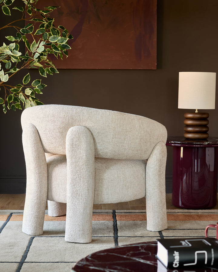 Anya accent chair in natural fabric with curved arms and cylindrical legs, displayed in a styled living room setting.