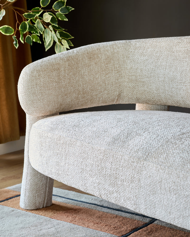 Anya accent sofa in natural fabric, shown at side angle highlighting curved armrests and cylindrical legs on patterned rug.
