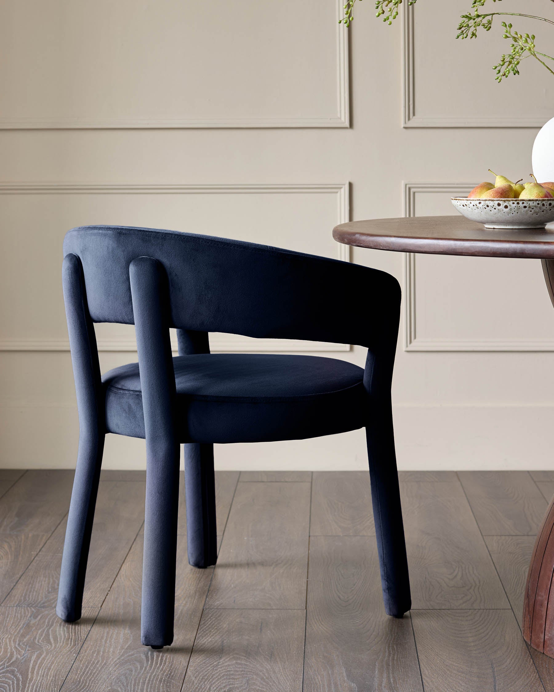 A stylish blue velvet dining chair sits beside a round table in a modern dining room. The chair has a curved back and sturdy legs.