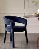 A navy blue velvet dining chair sits next to a round wooden table with a bowl of pears on it.