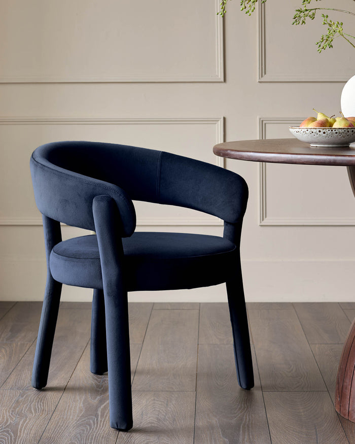 A navy blue velvet dining chair sits next to a round wooden table with a bowl of pears on it.