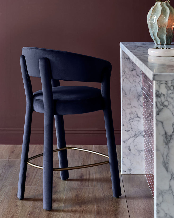 Dark navy velvet bar stool with curved backrest, brass footrest, and tapered legs in a styled interior setting.