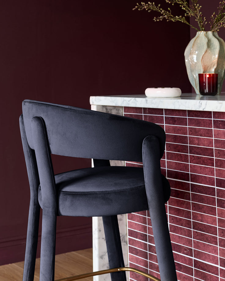 Anya bar stool in dark navy velvet with curved backrest and armrests, shown against burgundy wall.