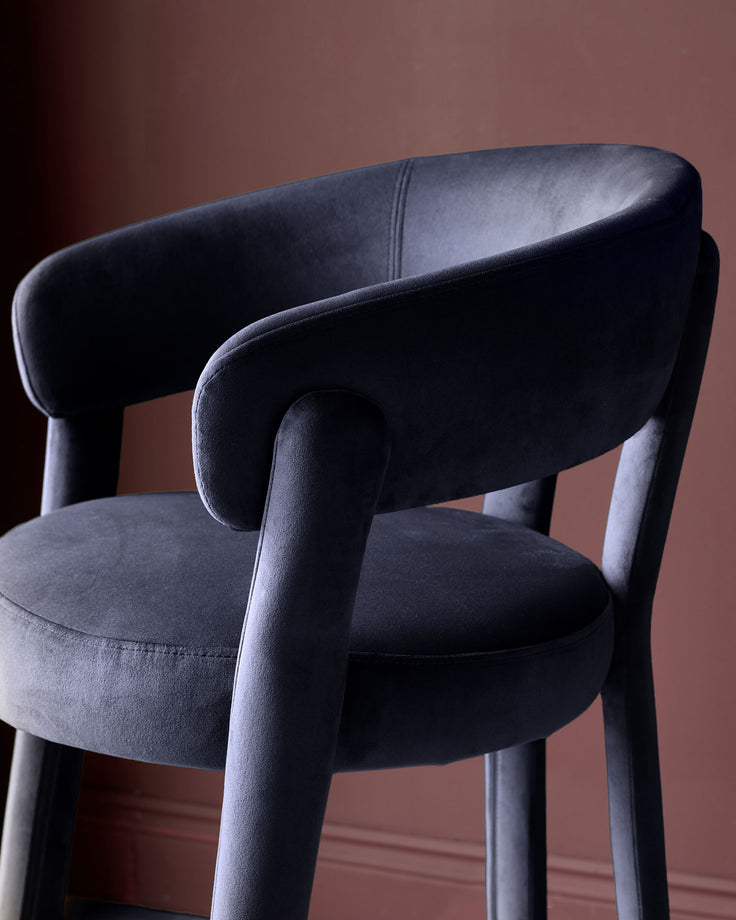 Anya bar stool in dark navy velvet with curved backrest and wooden legs, shown from side angle.